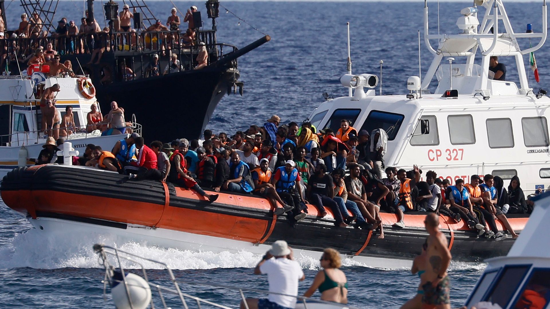 An Italian Coast Guard boat carries migrants as tourists on boat, foreground, watch, near the port of the Sicilian island of Lampedusa, southern Italy.